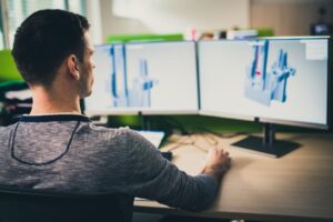 Over-the shoulder view of an engineer sitting at a desk and working on a 3D model of a machine.