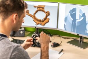 Engineer inspecting a machine part while making a 3D model on a computer.