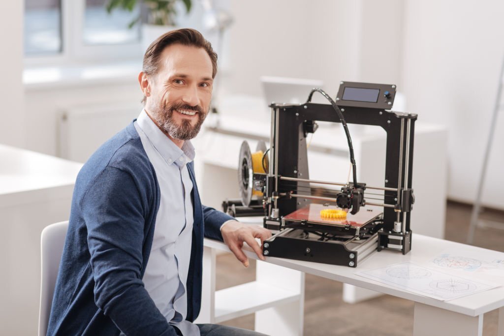 Technological advance. Handsome delighted positive man sitting near the 3d printer and smiling while looking at you